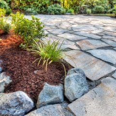 A garden with a paved stone pathway curving through it. The path, an exquisite example of estate maintenance, is lined with small plants and shrubs, bordered by a strip of mulch and large rocks. Sunlight filters through trees in the background, casting shadows on the ground.