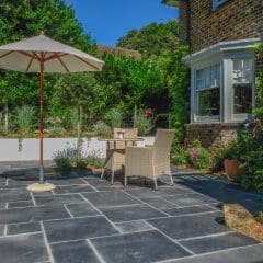 A sunlit patio with a table and two wicker chairs under a white umbrella, designed by an expert landscaper. The area is paved with large dark stones, surrounded by lush greenery and a brick house with white-framed windows.