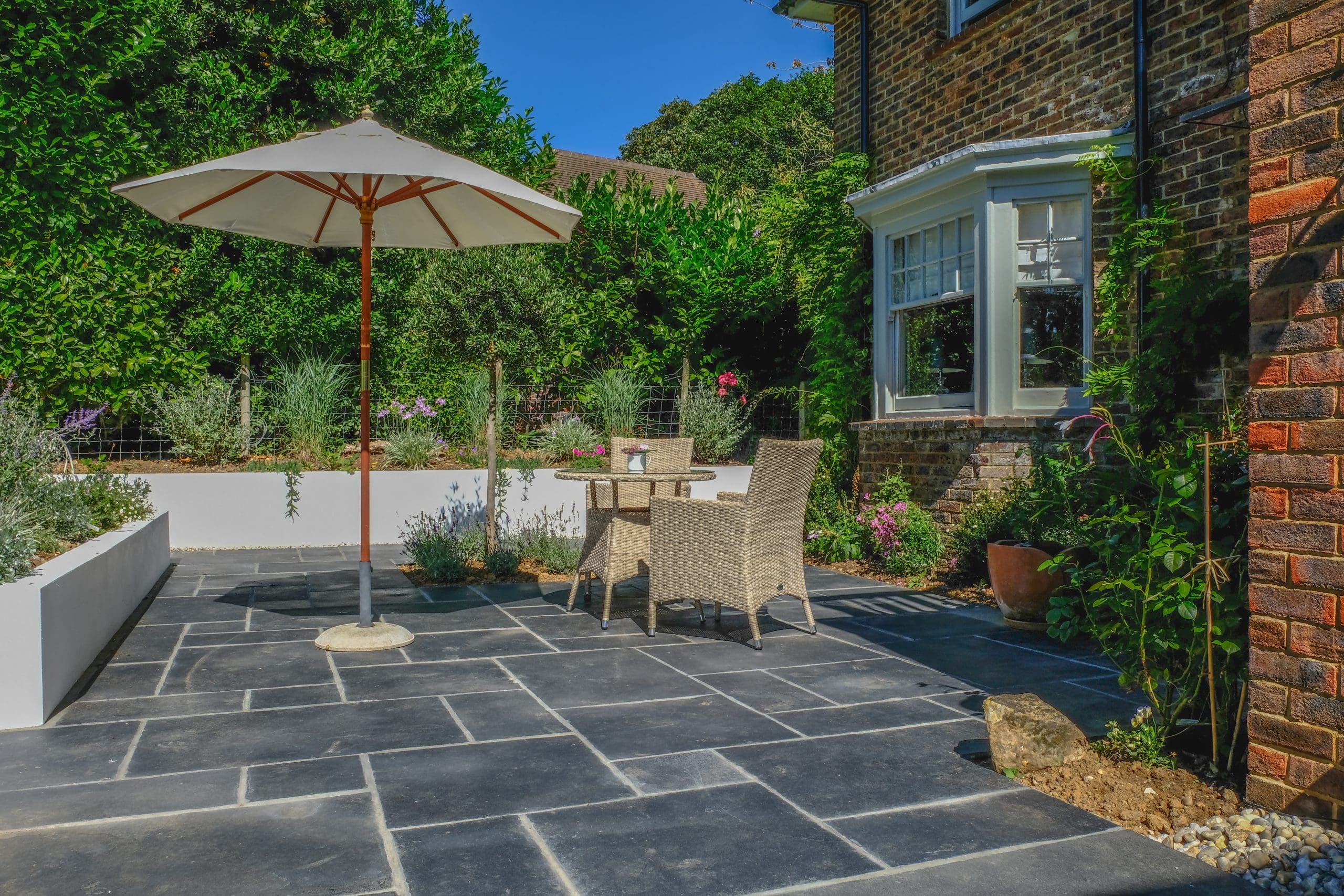 A sunlit patio with a table and two wicker chairs under a white umbrella, designed by an expert landscaper. The area is paved with large dark stones, surrounded by lush greenery and a brick house with white-framed windows.