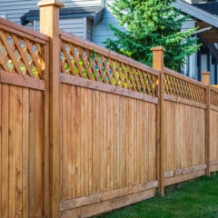 A wooden fence with a lattice design on top stretches along a suburban house, ideal for garden fencing. Made of light brown wood with evenly spaced vertical panels, it complements the tree and house visible in the background, perfect for estate maintenance.