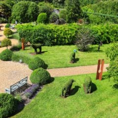 A picturesque garden featuring neatly trimmed hedges shaped like animals, a well-maintained lawn, sculptures, and gravel pathways. Benches are placed along the paths amidst elegant planters, surrounded by lush trees and shrubs under a clear sky. Estate maintenance ensures everything stays flawless.