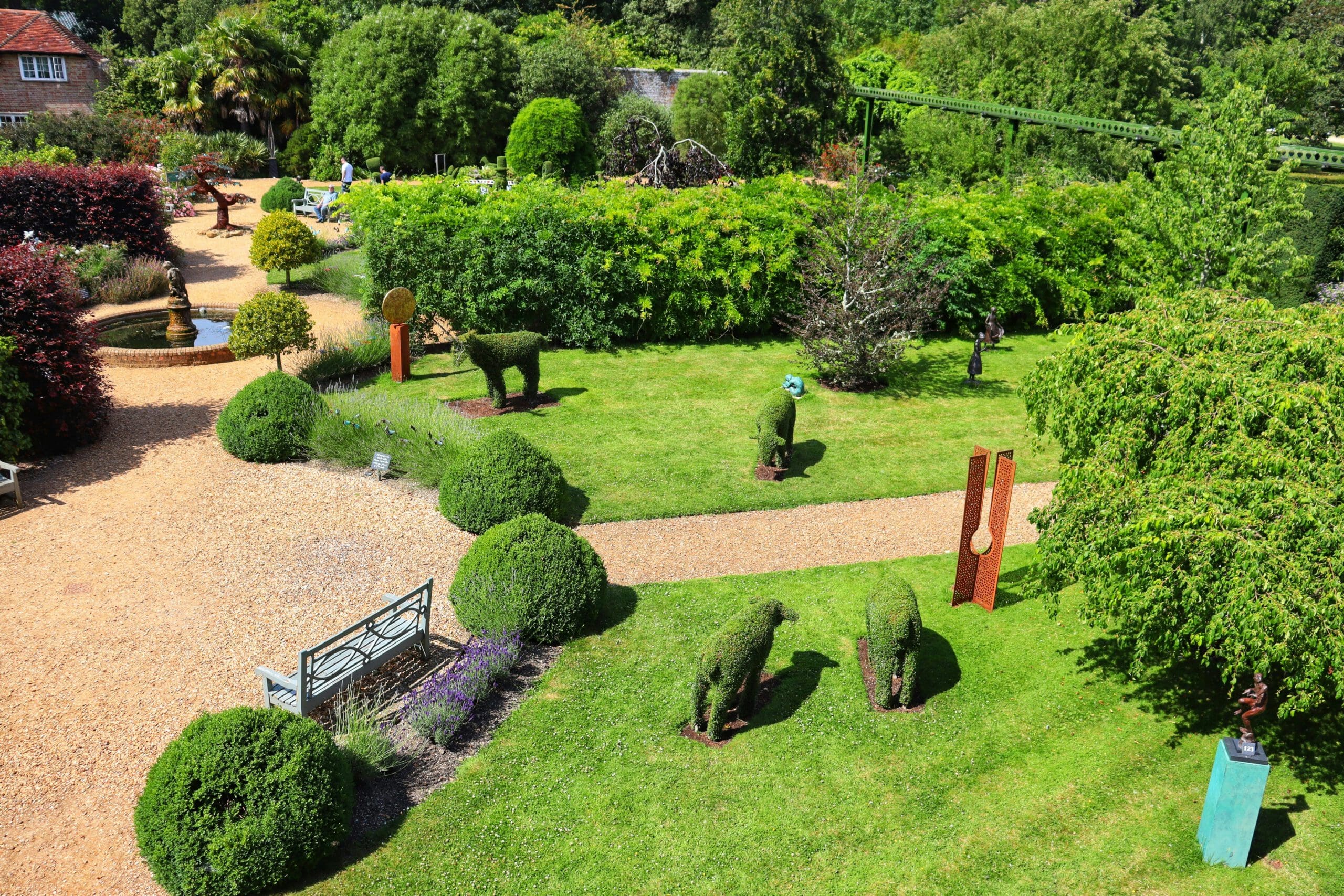 A picturesque garden featuring neatly trimmed hedges shaped like animals, a well-maintained lawn, sculptures, and gravel pathways. Benches are placed along the paths amidst elegant planters, surrounded by lush trees and shrubs under a clear sky. Estate maintenance ensures everything stays flawless.